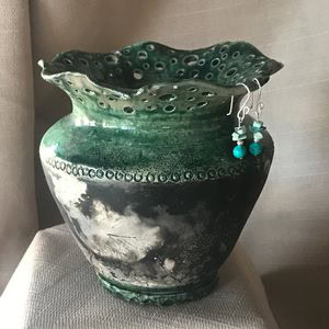 A green and black ceramic vase with a wavy, perforated rim sits on a surface; a pair of turquoise and white beaded earrings hang from the vases edge.