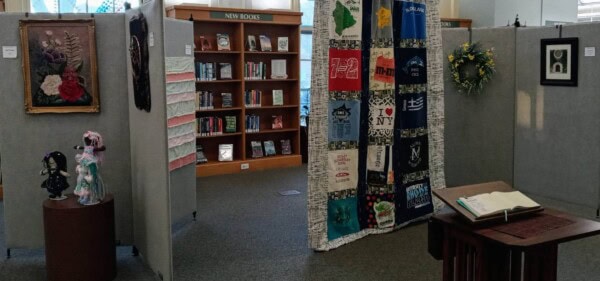 A library display area features quilts, framed art, and decorated dolls on gray panels. A shelf labeled New Books holds books in the background. A table in front displays an open guestbook and a floral wreath hangs on one panel.