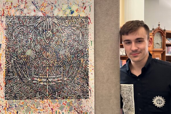 A young man in a black shirt with an abstract white design stands beside a colorful, intricate abstract painting with geometric patterns and splattered paint details, displayed indoors.