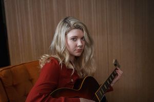 A young woman with long blonde hair wearing a red top sits on an orange chair and plays an acoustic guitar, looking toward the camera against a wooden panel background.