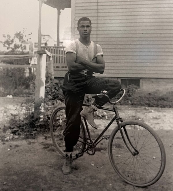 A young man stands confidently with arms crossed, one foot on the pedal of a vintage bicycle. He wears a tucked-in t-shirt, high-waisted pants, and boots. A house and porch appear blurred in the background.