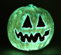 A pumpkin painted with glowing green paint, carved with triangular eyes and a jagged, toothy mouth, resembling a traditional jack-o’-lantern, set against a dark background.