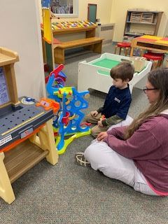 A young boy and an adult woman sit on the floor in a playroom, interacting with a colorful marble run toy. Various other toys and play equipment are visible in the background.