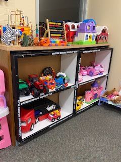 A variety of colorful childrens toys, including cars, trucks, a dollhouse, and an abacus, are neatly arranged on and inside a wooden shelf in a room with carpeted flooring.
