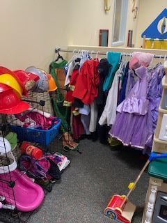 A small room with children’s dress-up clothes hanging on racks, including costumes like a princess dress and superhero outfits. Toy helmets, hats, and accessories are stored in bins and on shelves nearby.