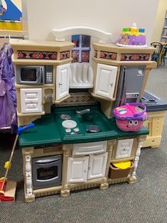 A plastic childrens play kitchen set with a green countertop, stove, sink, microwave, oven, fridge, and cabinets. A pink basket toy sits on the counter, and a play broom leans against the set.