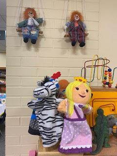 Two hand puppets—a zebra and a princess—sit on a shelf in front of colorful toys. Above them, two clown dolls in bright outfits are seated on swings attached to a cream brick wall.