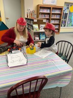 An adult and a child, both wearing red plastic firefighter hats, sit at a table with coloring pages and crayons in a library. Bookshelves and colorful decorations are visible in the background.