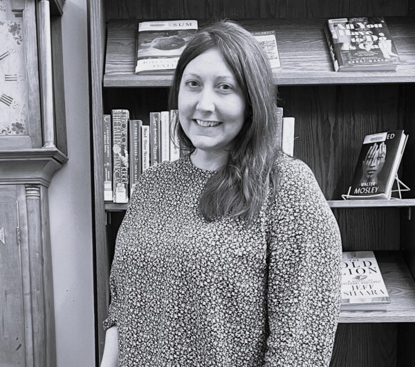 A woman with long hair, wearing a patterned top, smiles while standing in front of a bookshelf filled with books in a library or bookstore setting.