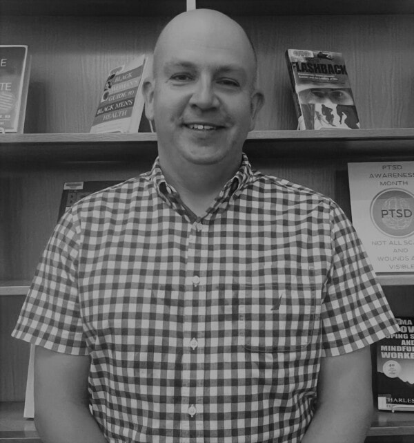 A man in a checkered short-sleeve shirt stands smiling in front of a bookshelf displaying books and a PTSD Awareness Month sign. The photo is in black and white.
