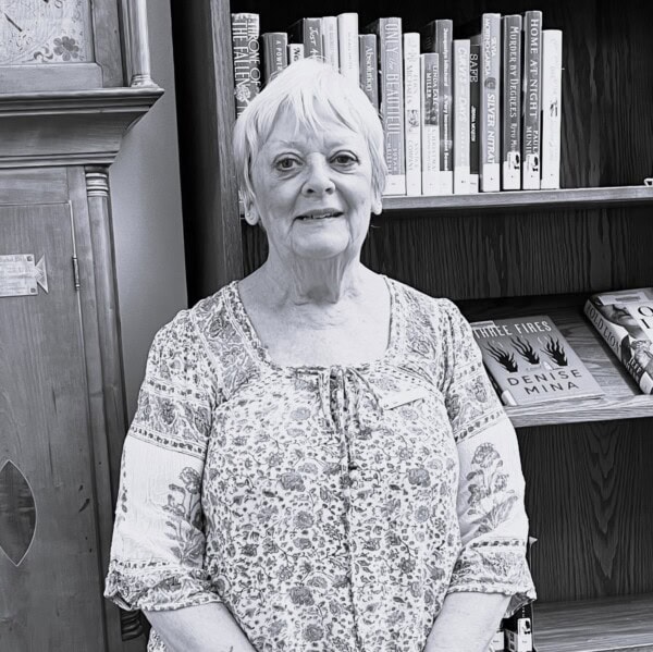 An older woman with short, light hair and a patterned blouse stands smiling in front of a bookshelf filled with books. The photo is in black and white.