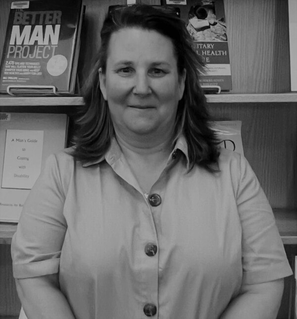 A woman with shoulder-length hair and a buttoned shirt stands in front of a bookshelf holding various books, including Better Man Project. The photo is in black and white.