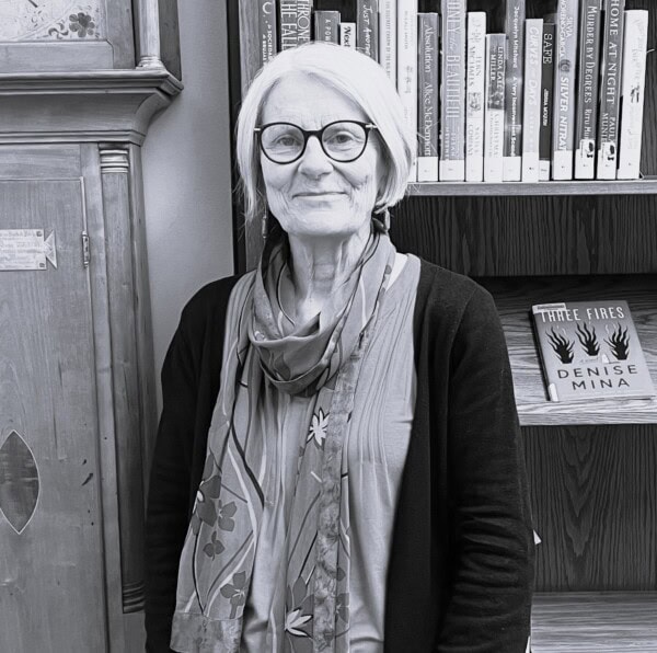 An older woman with glasses and a patterned scarf stands smiling in front of bookshelves filled with books, including “Three Fires” by Denise Mina. The image is in black and white.