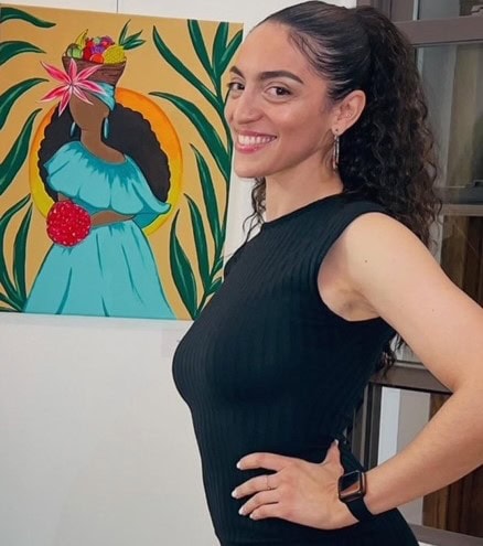 A woman with curly hair in a ponytail, wearing a fitted black dress and a smartwatch, smiles while standing sideways in front of a colorful painting of a woman in a blue dress holding fruit.