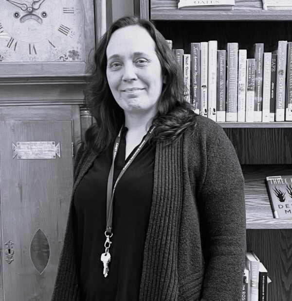 A woman with wavy dark hair, wearing a cardigan and a lanyard with keys, stands in front of a bookshelf filled with books and a large antique clock. The image is in black and white.