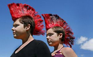 Two people with short brown hair and tall, spiked red mohawks stand side by side outdoors under a bright blue sky with a few clouds.