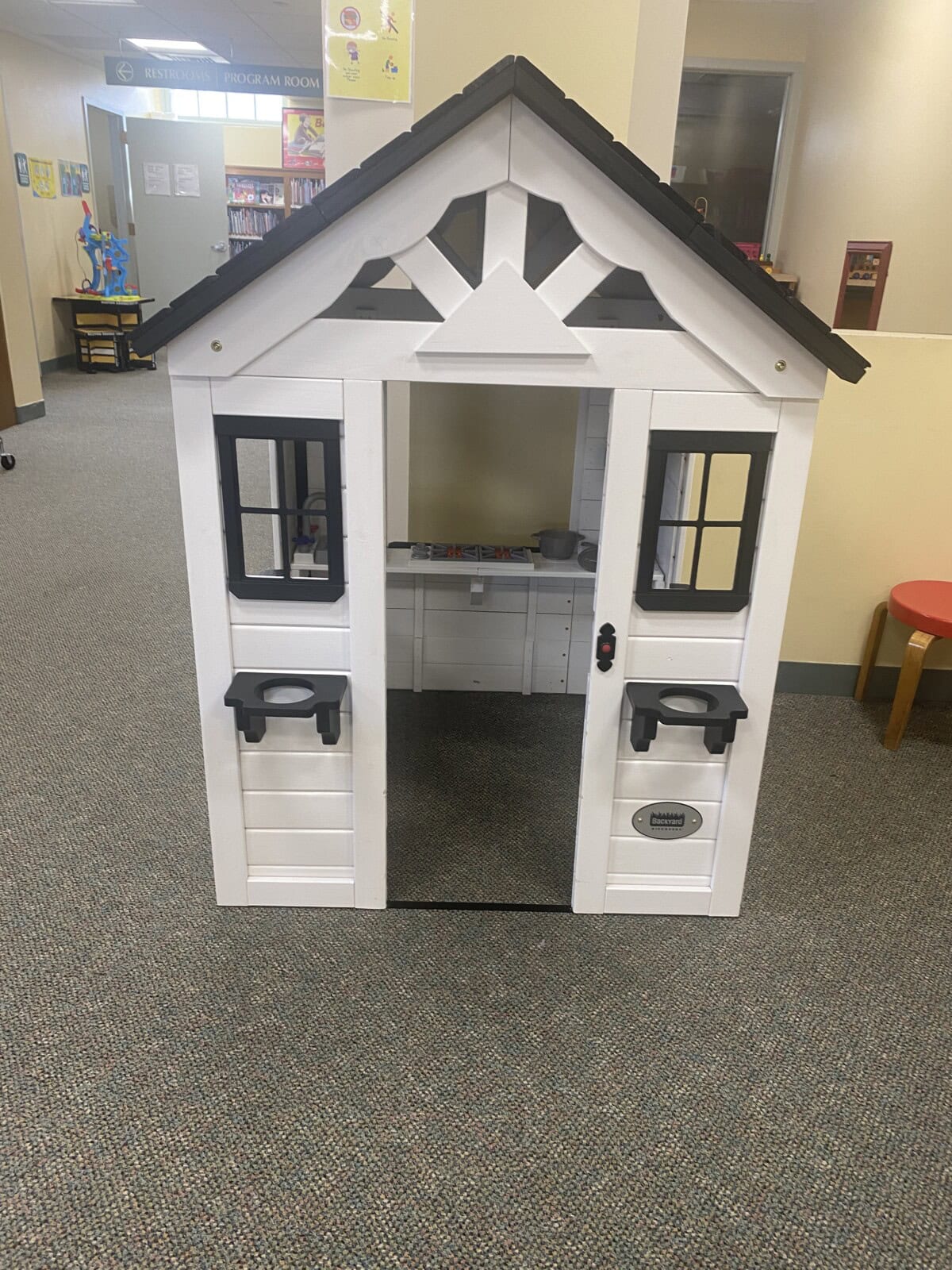 A small white playhouse with black trim and open door sits on a carpeted floor in a library or classroom, with a red stool and bookshelves visible in the background.