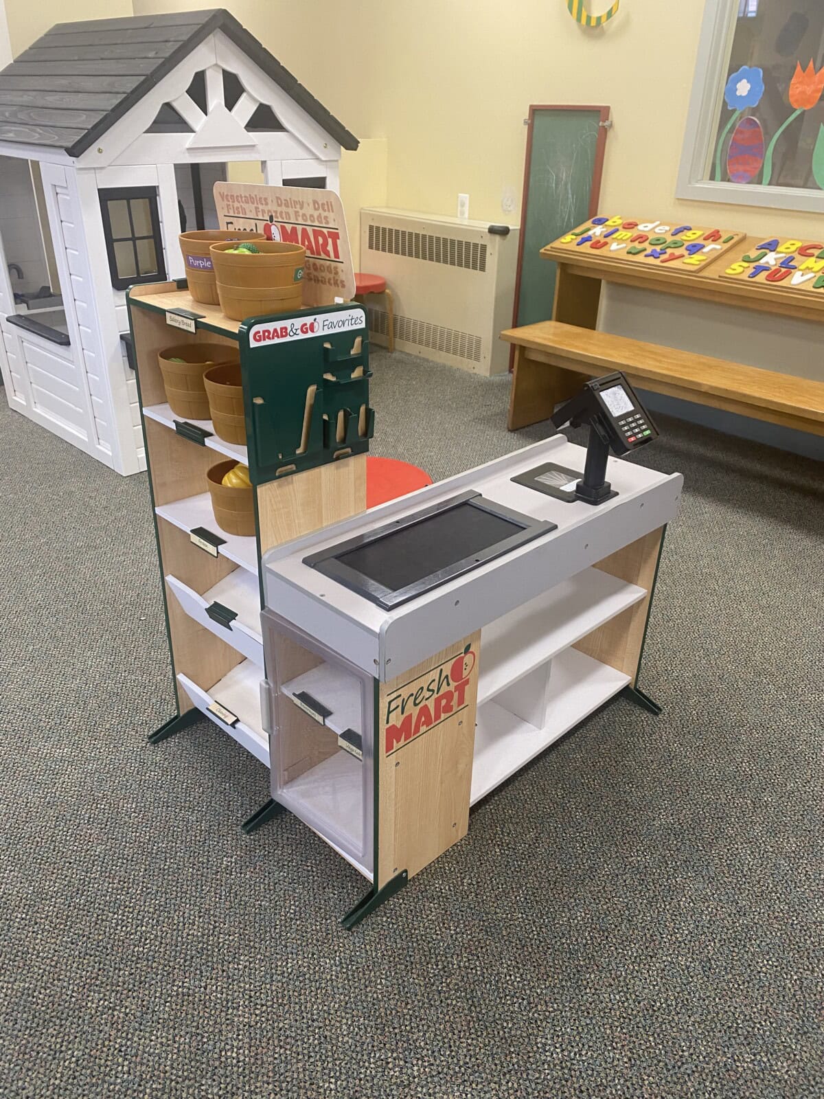 A childrens play grocery store checkout with a toy cash register, scanner, and shelves, sits in a playroom. A small white playhouse and wooden abacus are visible in the background.