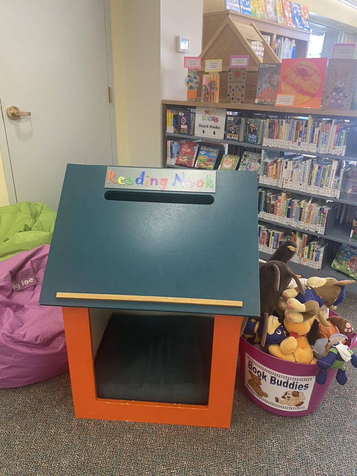 A small reading nook for children sits next to a purple beanbag, a basket of stuffed animals, and shelves filled with books in a library’s children’s section. The nook has a sign labeled Reading Nook.