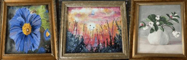Three framed paintings: a close-up of a blue flower with green leaves, a sunset with a white bird above wildflowers, and a white vase holding branches with white flowers and green leaves.