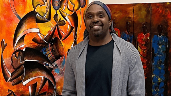 A smiling man in a gray cardigan and black shirt stands in front of a colorful abstract painting with vivid red, orange, and yellow hues and figures in the background.