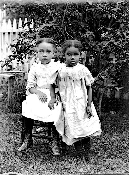 Two young girls in dresses pose for a black-and-white portrait outdoors. One is seated in a chair, and the other stands beside her. A leafy bush and a wooden picket fence are visible in the background.